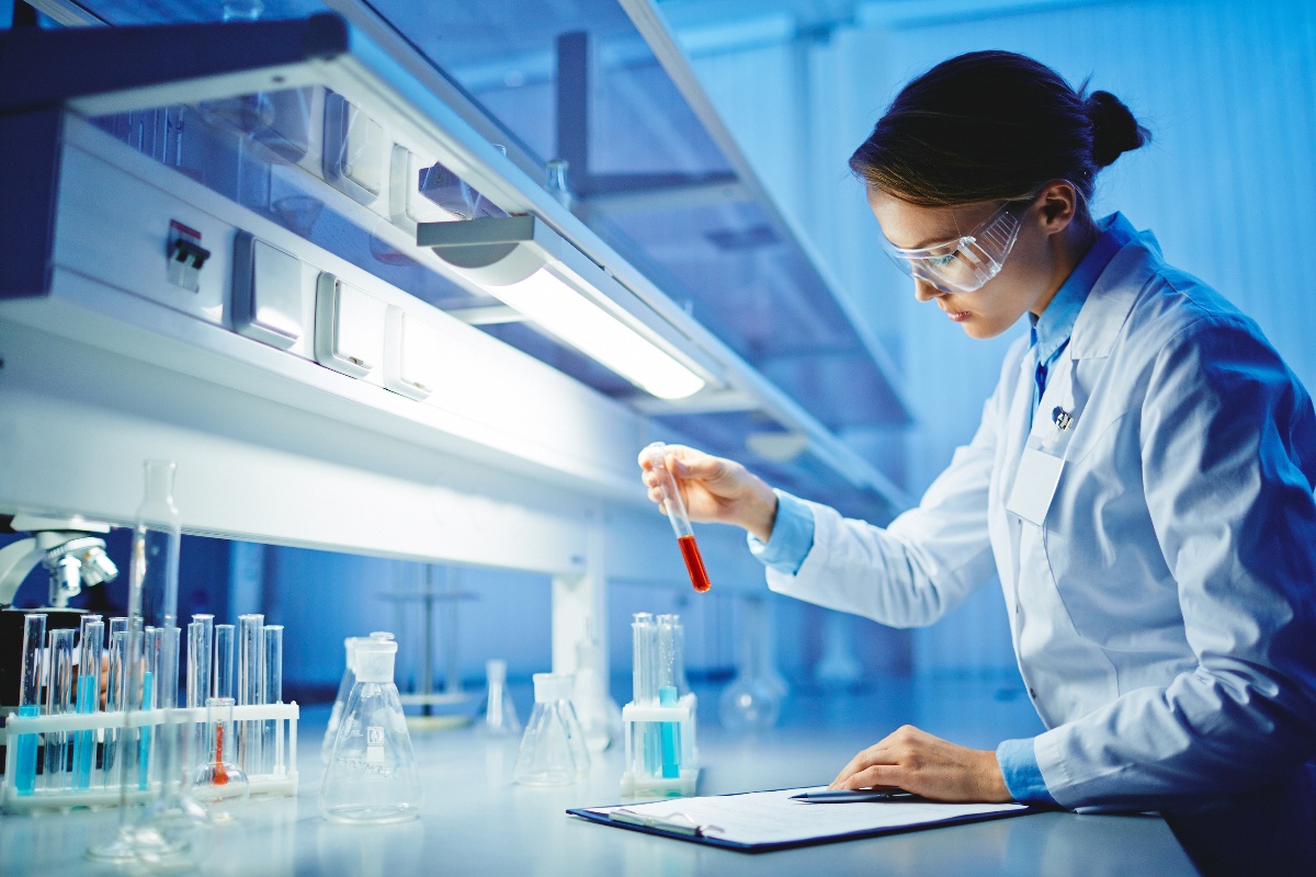 A scientist wearing safety goggles and a lab coat examines a test tube with red liquid in a laboratory, with various glassware and equipment on the bench.