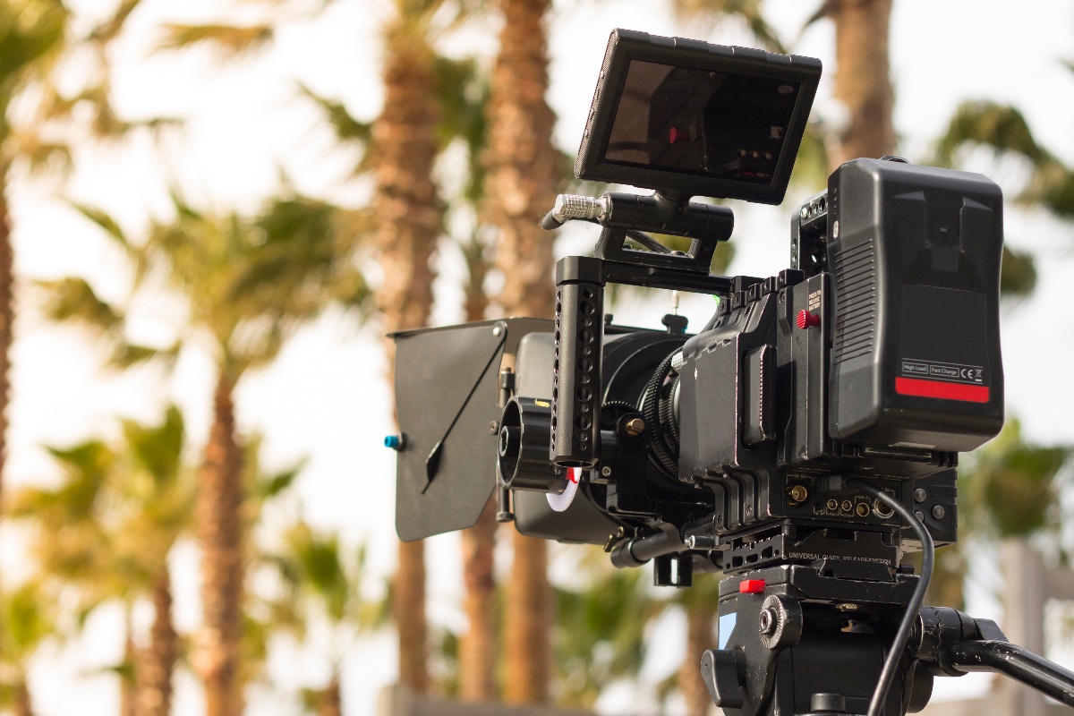 Close-up of a professional video camera set up outdoors, with palm trees blurred in the background.