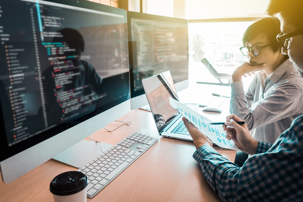 Two people work on a code review at a desk with multiple computer monitors displaying code and data graphs. One person points to a printed sheet, while the other looks at the screen thoughtfully.