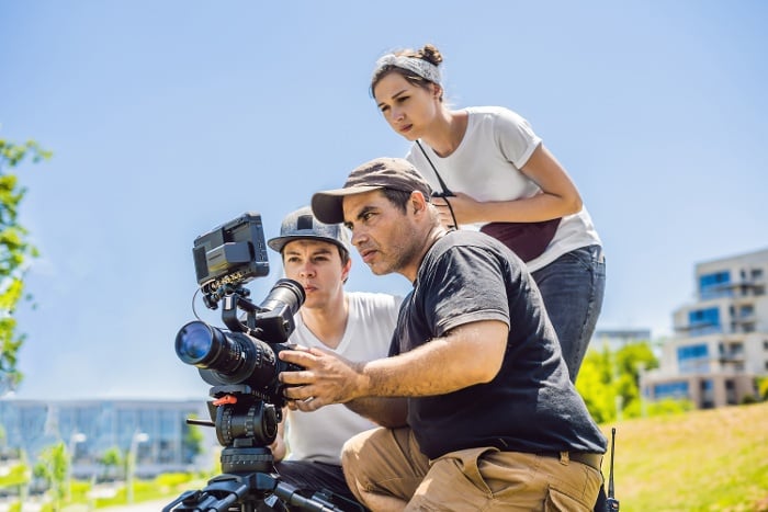 Three people operating a video camera outdoors, focusing intently on the camera screen with a sunny city park in the background.