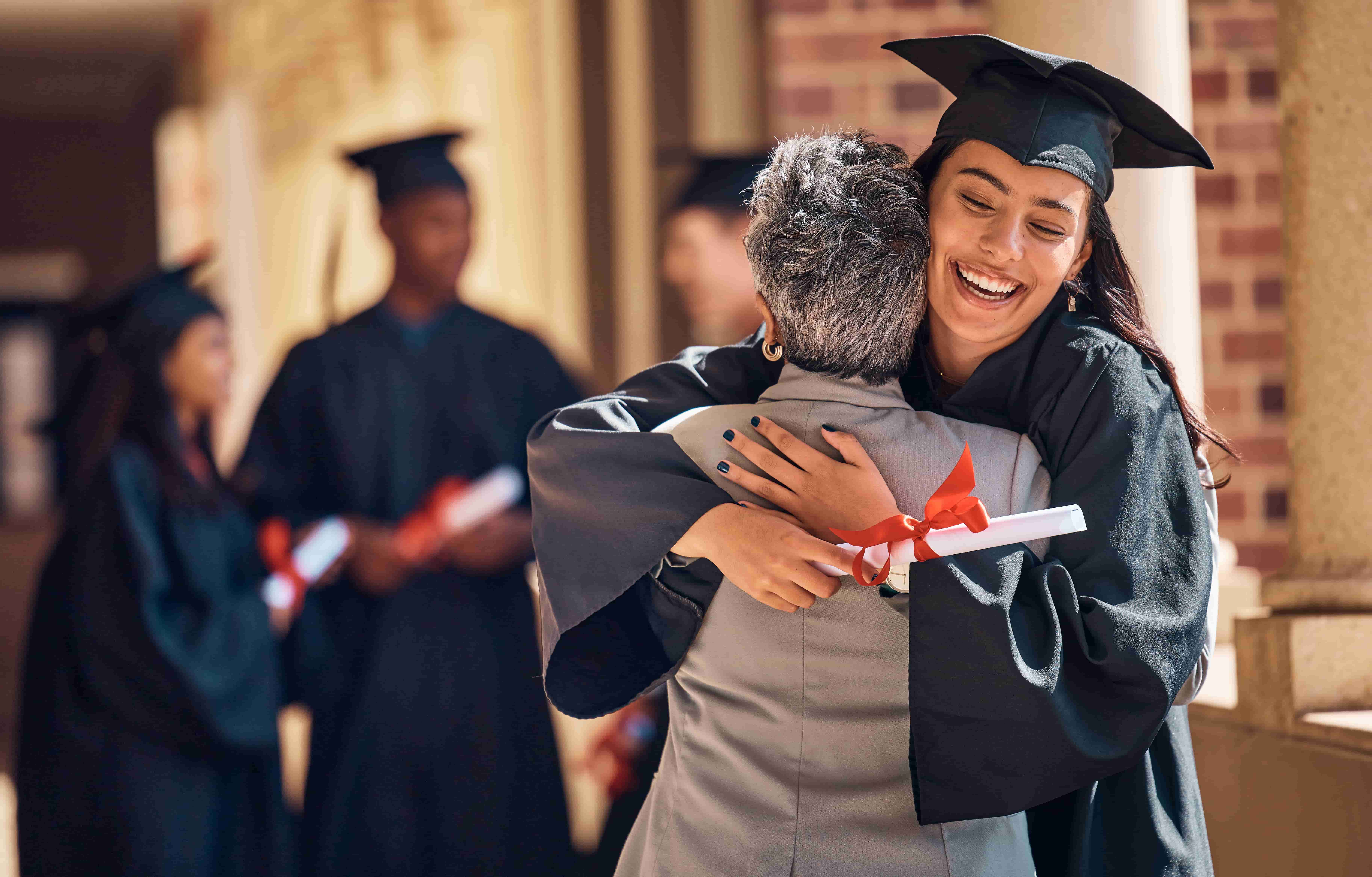 graduate in a cap and gown hugs an older person while holding a diploma. Other graduates can be seen in the background.