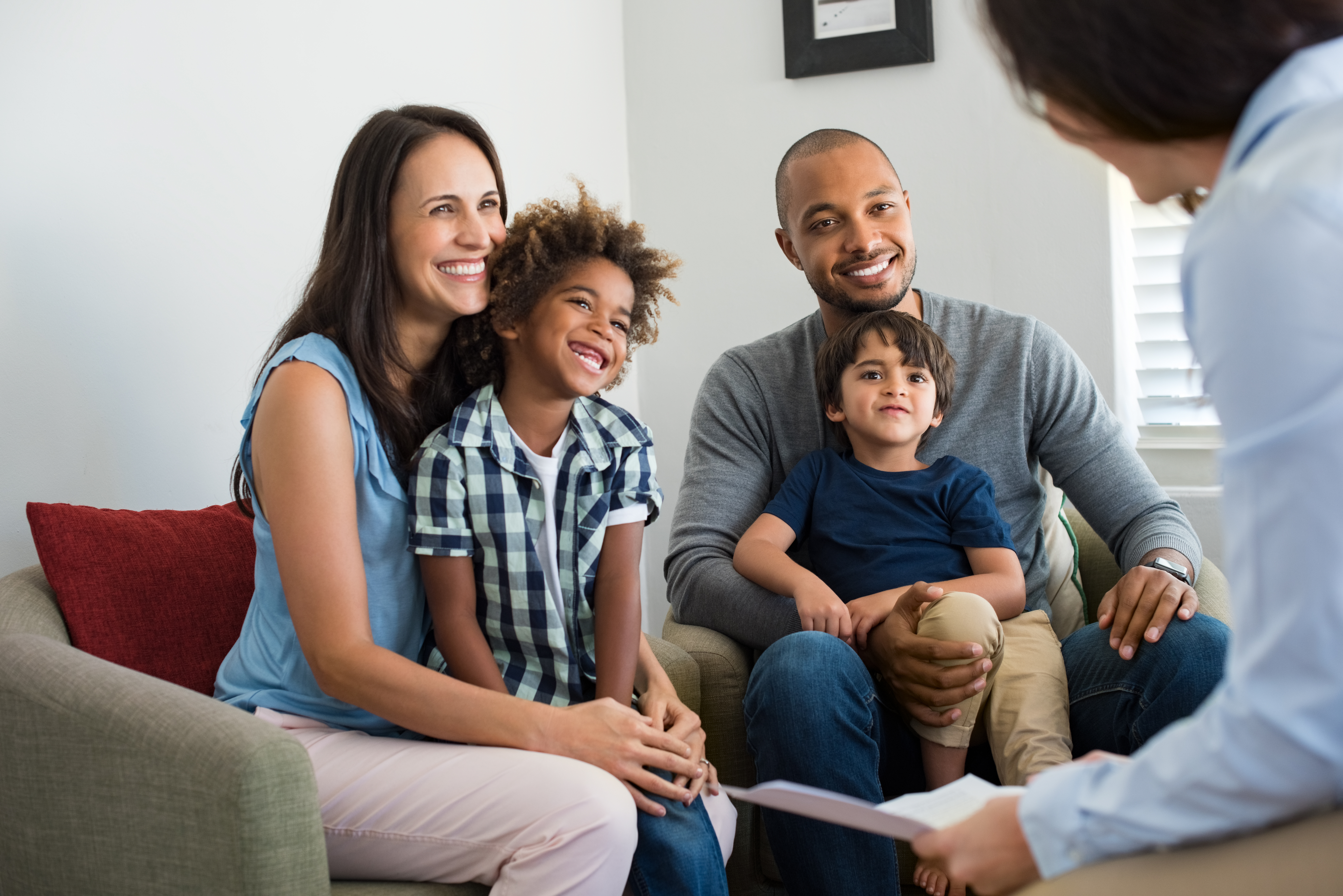 A smiling family of four sits together on a couch, talking to a woman holding papers in an indoor setting.