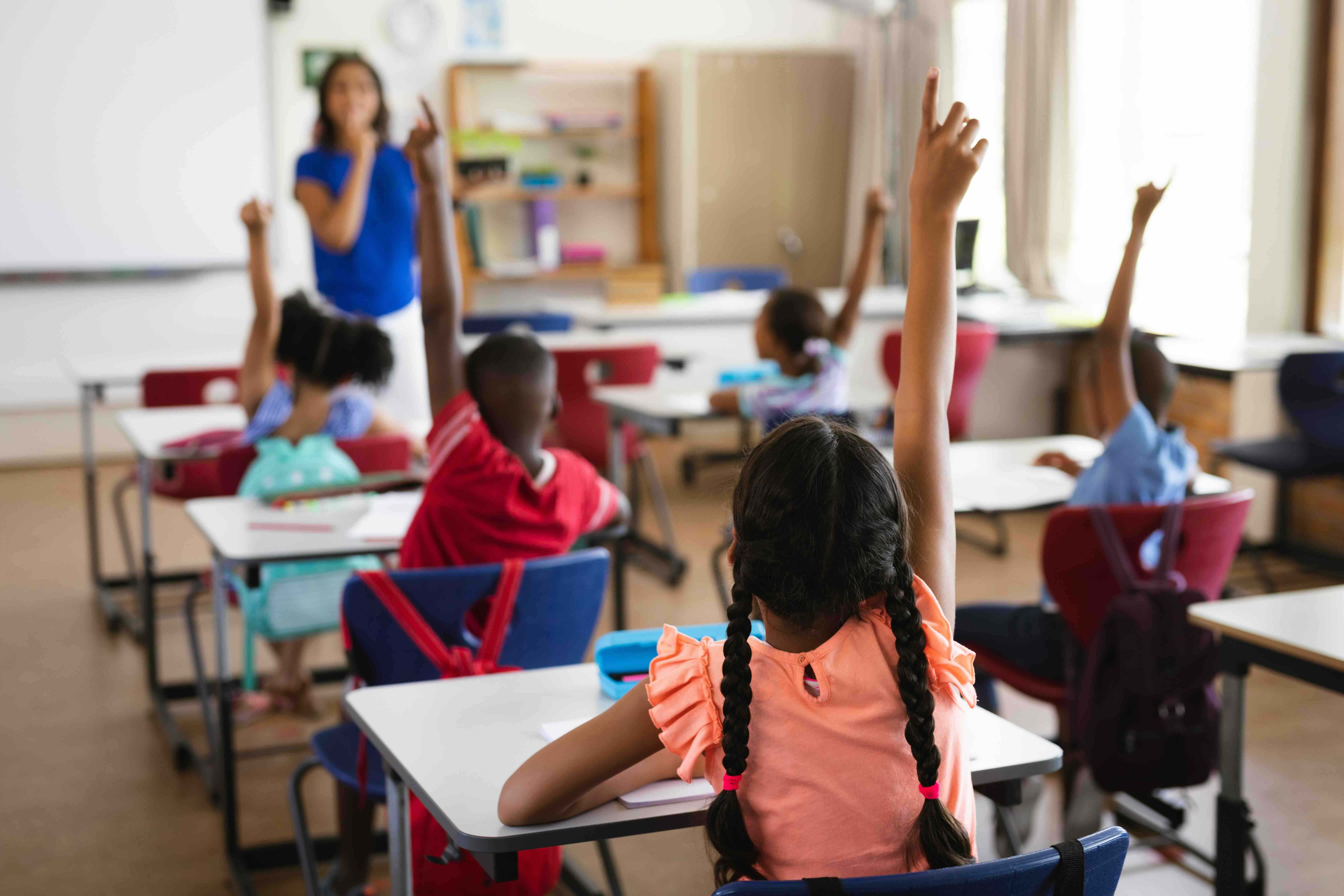 Students seated at desks in a classroom raise their hands as a teacher stands at the front of the room.
