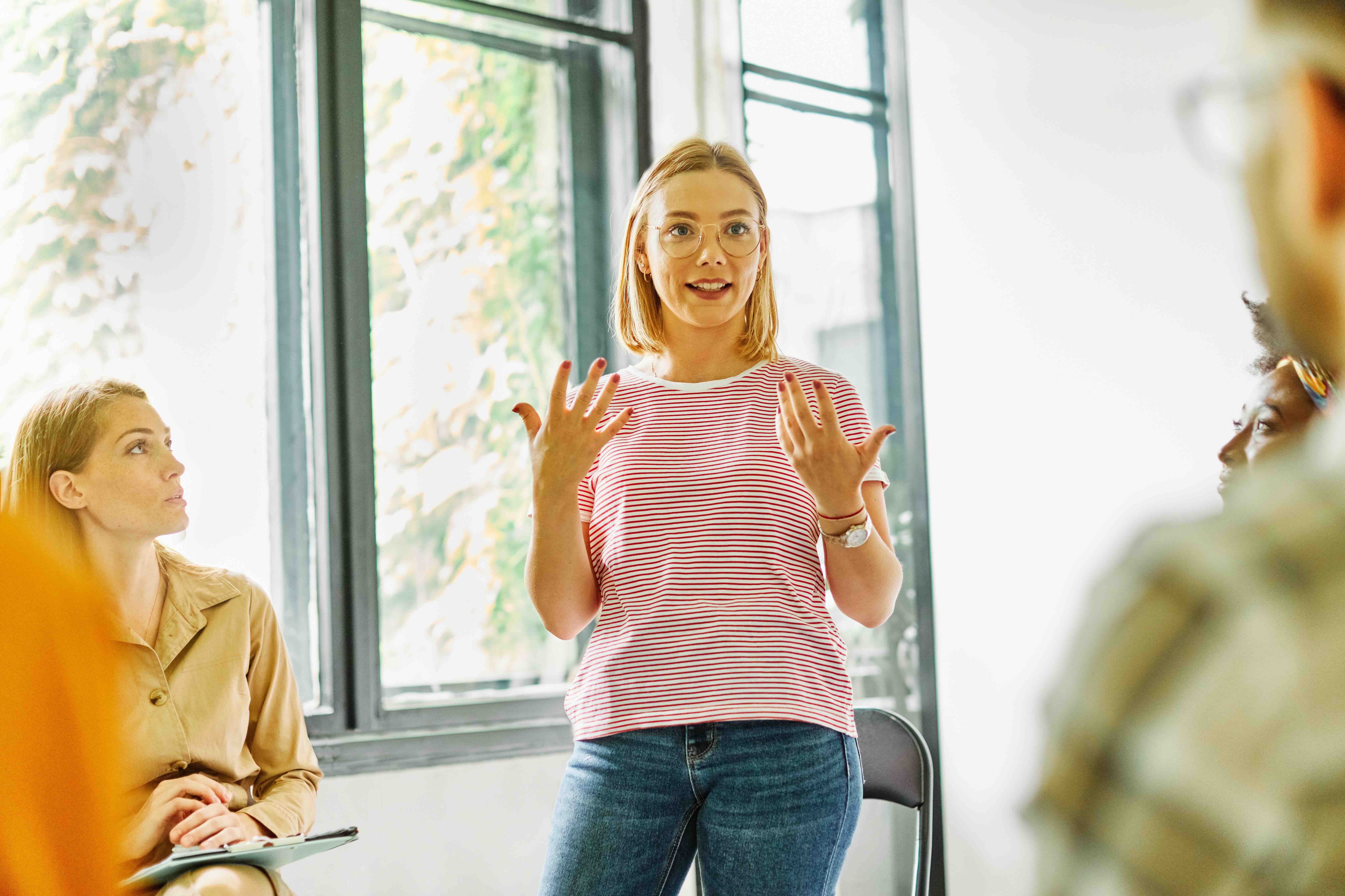 A woman in a striped shirt stands and speaks to a group in a bright room, while others sit and listen attentively.