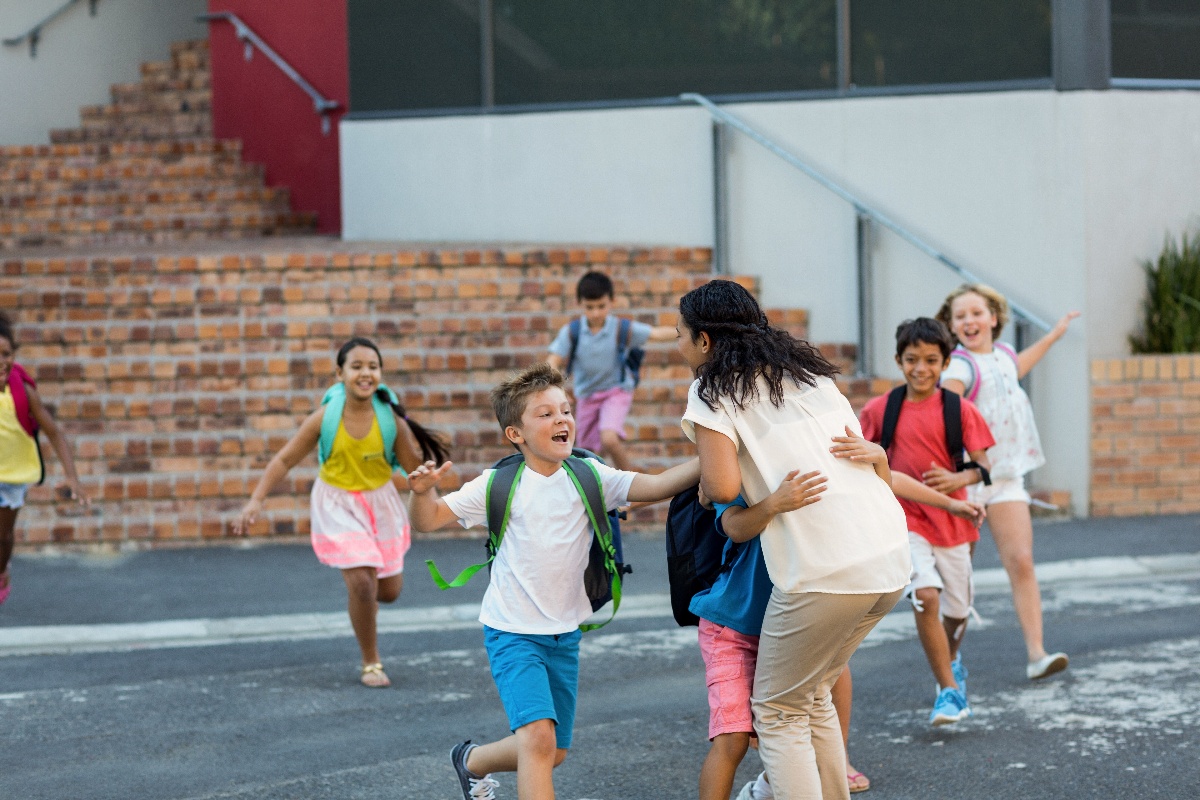 A group of children, wearing backpacks and smiling, run toward a woman for hugs outside a building with brick steps.