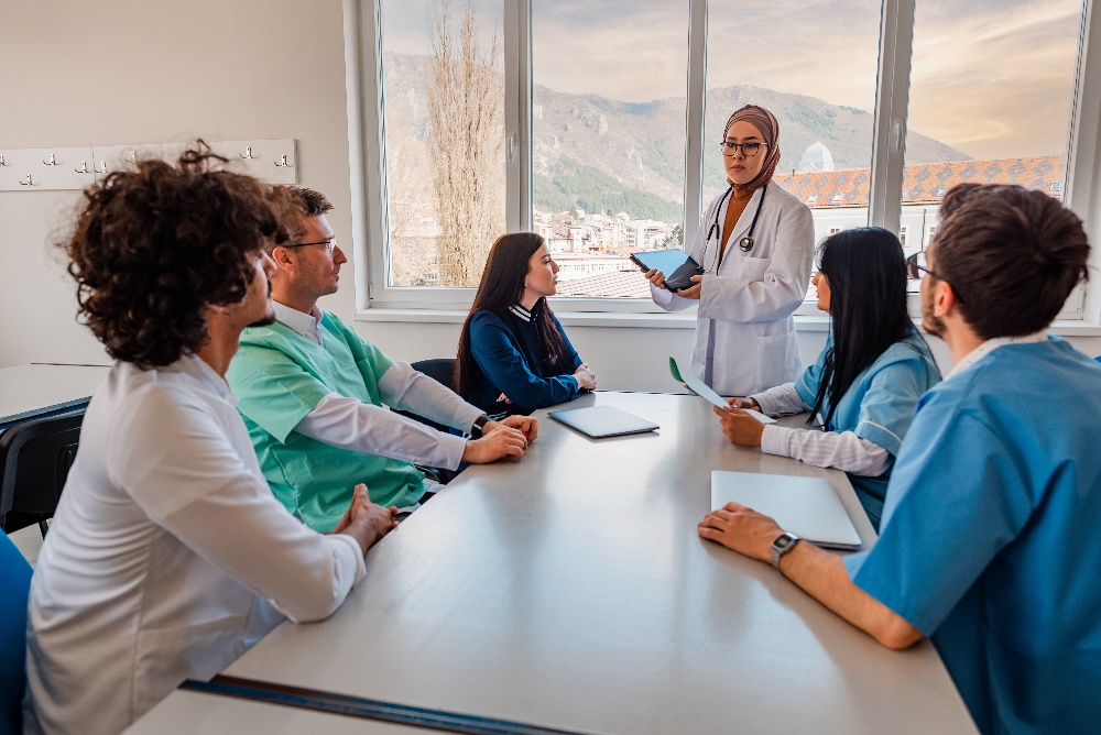 A doctor stands and presents to five seated colleagues in a bright conference room with a large window and mountain views.