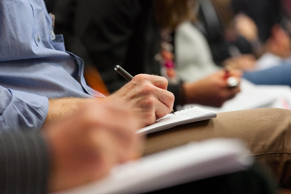 People are seated and taking notes with pens and notebooks during a meeting or conference.