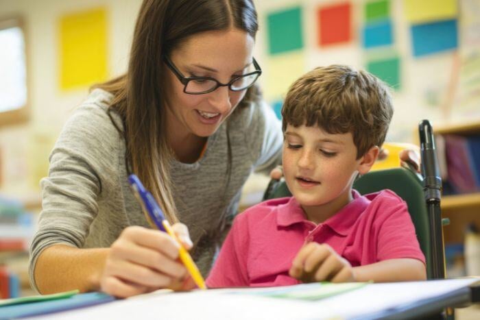 A woman with glasses is helping a young boy with his work at a table in a classroom setting.