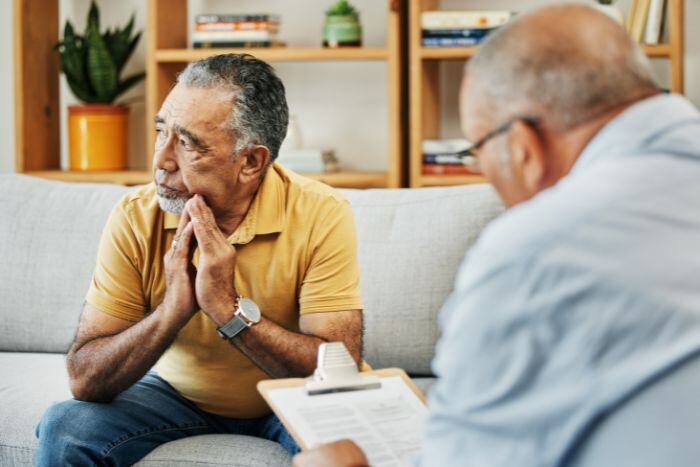 Two men are sitting facing each other in a living room. One man is in a yellow shirt, looking contemplative, while the other holds a clipboard and appears to be taking notes.